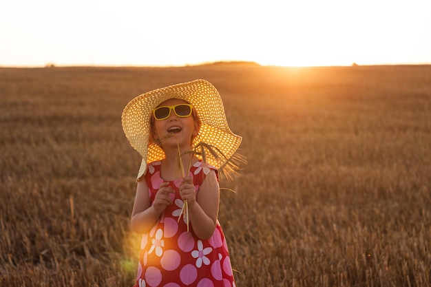 Adorable niña con sombrero de paja y vestido rosa de verano en campo de trigo Niño con cabello largo y rubio en la agricultura al atardecer