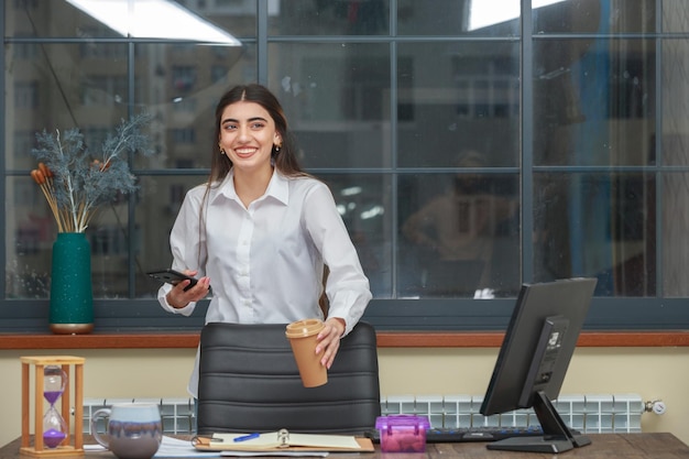 Adorable niña sosteniendo una taza de café y un teléfono mientras sonríe a la cámara