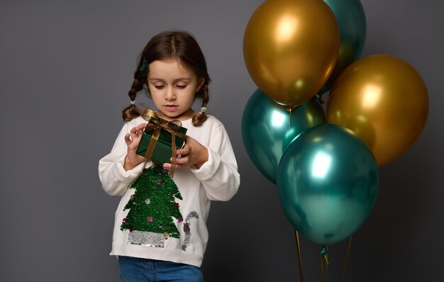 Adorable niña sostiene su regalo de Navidad en papel de regalo verde brillante y cinta dorada, se encuentra cerca de hermosos globos de aire verdes brillantes metálicos y dorados, aislados sobre fondo gris, espacio de copia