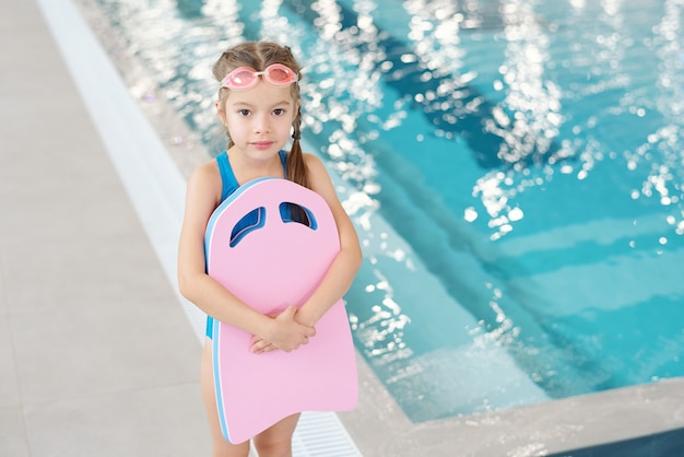 Adorable niña en traje de baño y gafas con tabla de natación mientras está de pie frente a la cámara contra la piscina en el centro deportivo