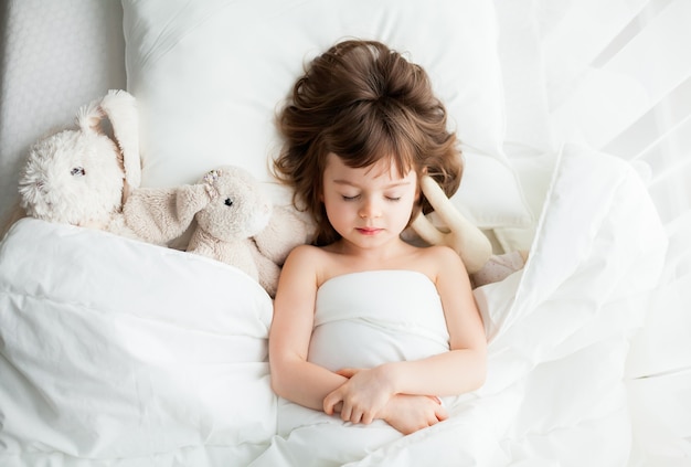 Adorable niña tranquila durmiendo en la cama blanca con juguetes de conejo cerca de ella