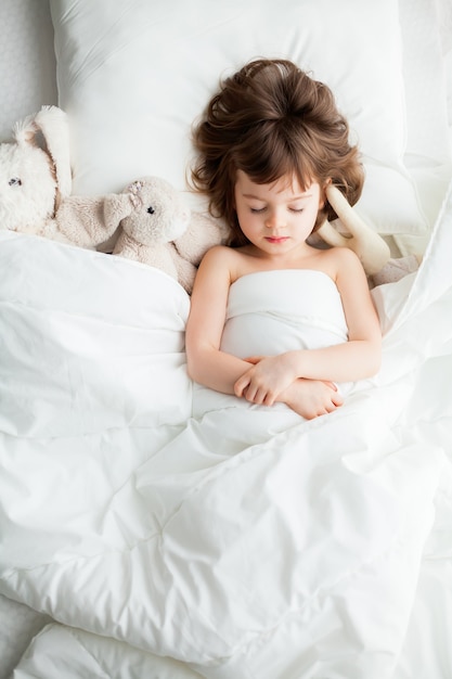 Adorable niña tranquila durmiendo en la cama blanca con juguetes de conejo