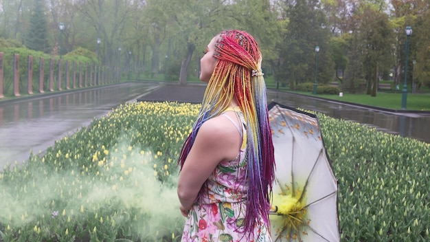 Adorable niña con un vestido con estampado floral con trenzas de arcoíris y maquillaje. Danzas con un paraguas escondido en humo amarillo con el telón de fondo de la cama de flores con tulipanes