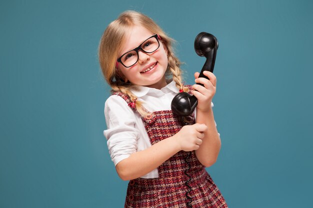 Adorable niña de vestido rojo, camisa blanca y gafas tiene teléfono auricular