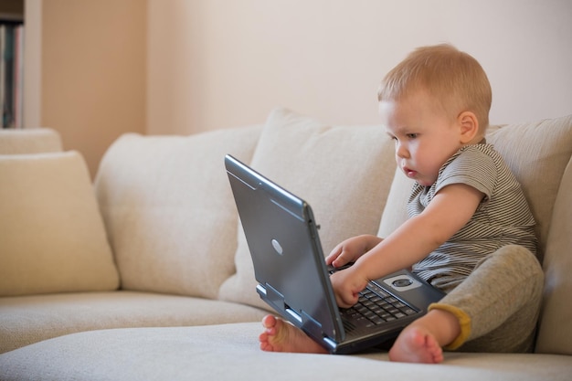 Adorable niñito sentado en un sofá y jugando con una computadora de juguete