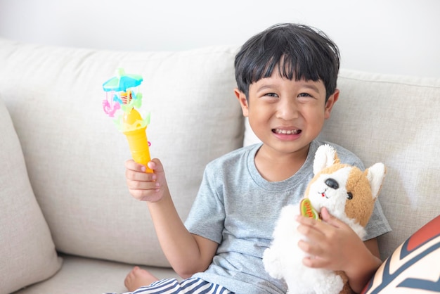 Adorable niño asiático feliz mirando a la cámara con una camisa gris y pantalones cortos a rayas azules y blancas sentado en un sofá crema sonríe feliz La muñeca del perro de la mano izquierda y la mano derecha sostiene un juguete