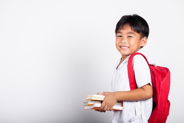 Adorable niño asiático sonriendo feliz vistiendo uniforme tailandés de estudiante pantalones rojos libros de pie para estudiar listos para la escuela aislados sobre fondo blanco Retrato niños pequeños niño preescolar Regreso a la escuela