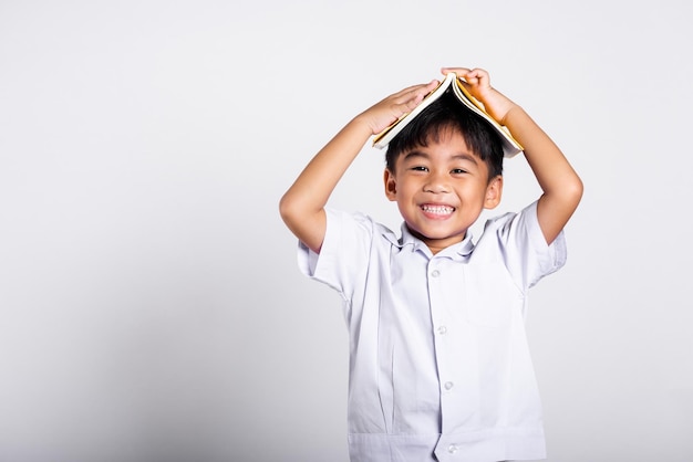 Un adorable niño asiático sonriendo feliz vistiendo un uniforme tailandés de estudiante con pantalones rojos de pie sosteniendo un libro sobre la cabeza como un techo