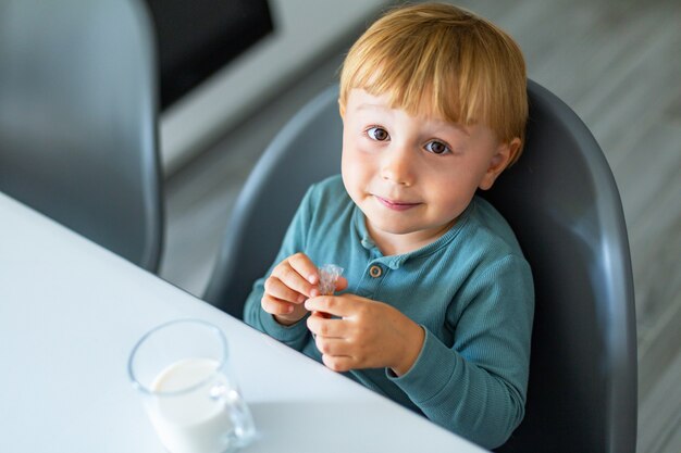 Adorable niño bebiendo leche o yogur y comiendo caramelo desayunando en la cocina por la mañana