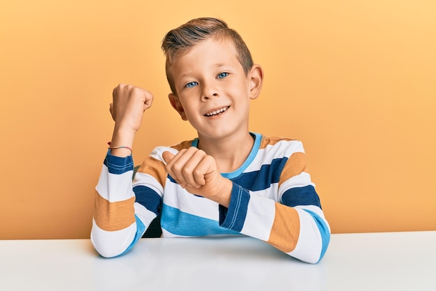 Adorable niño caucásico con ropa informal sentado en la mesa apuntando hacia atrás con la mano y los pulgares hacia arriba, sonriendo confiado