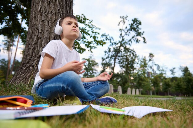 Foto adorable niño escolar se siente agotado y cansado después de la escuela y la tarea, se sienta en posición de loto y medita con auriculares inalámbricos en la cabeza. libros de trabajo y útiles escolares tirado en el césped