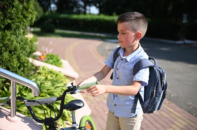 Adorable niño estacionando su bicicleta en la entrada de la escuela. Chico guapo regresando a la escuela