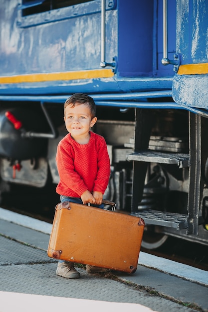 Adorable niño niño vestido con suéter rojo en una estación de tren cerca de tren con maleta marrón vieja retro. Listo para vacaciones. Viajero joven en la plataforma.