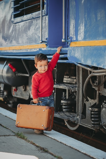 Adorable niño niño vestido con suéter rojo en una estación de tren cerca de tren con maleta marrón vieja retro. Listo para vacaciones. Viajero joven en la plataforma.