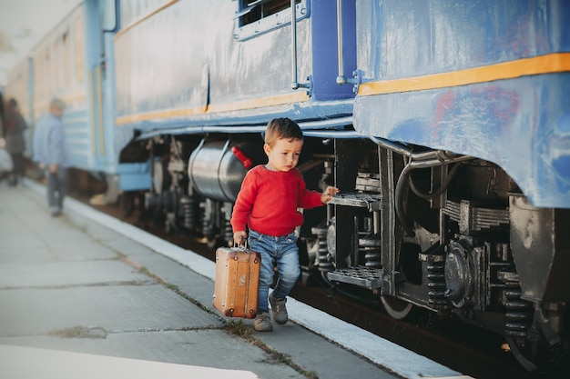 Adorable niño niño vestido con suéter rojo en una estación de tren cerca de tren con maleta marrón vieja retro. Listo para vacaciones. Viajero joven en la plataforma.