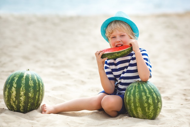 Adorable niño en la orilla del mar comiendo jugosa sandía. Niño alegre en verano en la playa. Niño pequeño lindo al aire libre