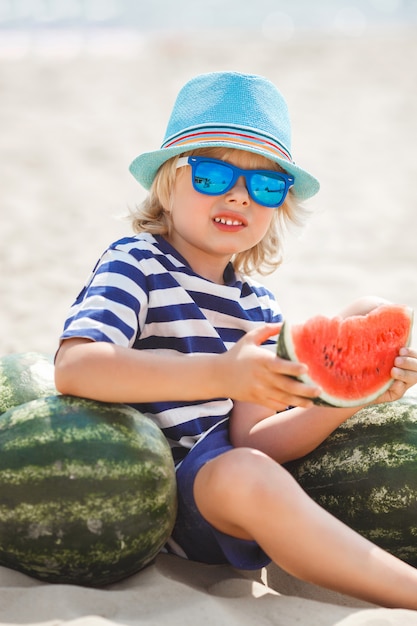 Foto adorable niño en la orilla del mar comiendo jugosa sandía. niño alegre en verano en la playa. niño pequeño lindo al aire libre