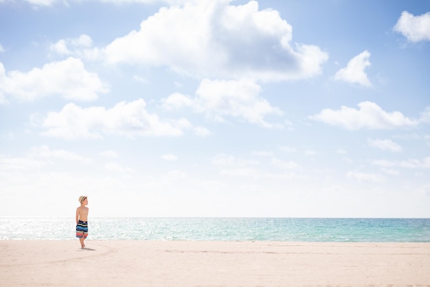 Adorable niño parado en la playa de arena junto al océano y mirando la hermosa vista de verano
