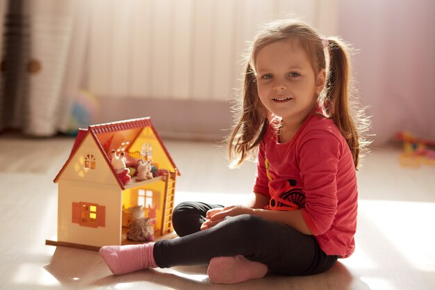 Adorable niño pequeño con dos coletas, niña de edad preescolar jugando con una casa de juguetes sentada en el piso en una habitación soleada, pasando tiempo en casa