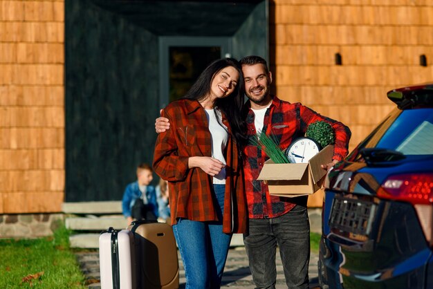 Adorable pareja de enamorados sonriendo y sintiendo felicidad cuando está de pie cerca de la casa moderna con caja de cartón con reloj y plantas verdes durante la reubicación a la nueva casa.