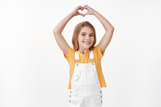 Adorable tierna jovencita con cabello rubio, use camiseta amarilla de verano, monos, levante las manos y muestre el signo del corazón