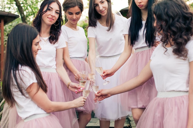 Adorables chicas de pie en un círculo con una joven novia, sonriendo, sosteniendo copas con champán. Despedida de soltera con hermosas novias en faldas de tul rosa