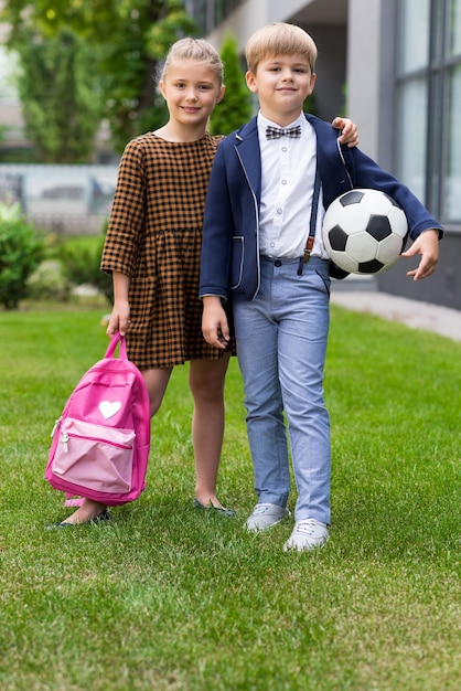 Adorables escolares sosteniendo mochila y pelota de fútbol mientras están de pie juntos cerca de la escuela