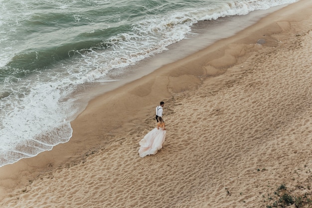 Adorables novios se toma de la mano caminando por la playa contra las olas del mar.