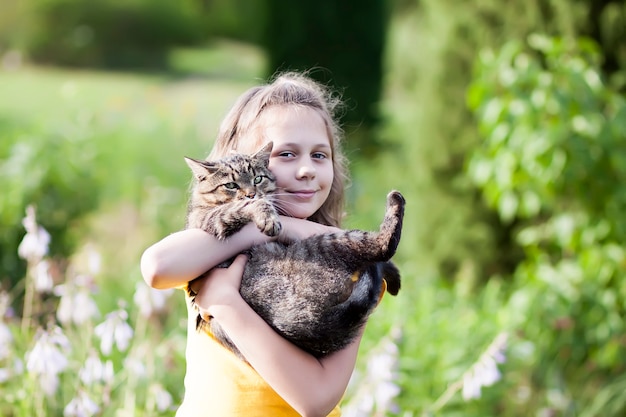 Adorale-Mädchen im gelben Kleid, das reizende Katze in ihren Händen hält. Kind und Haustier draußen am Sommertag.
