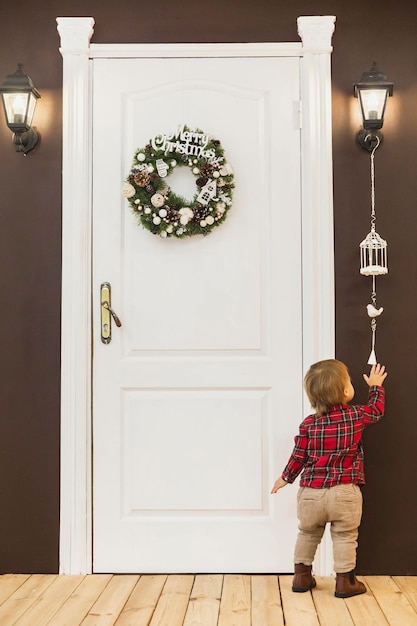 adorável bebê fica na porta da frente com uma guirlanda de natal