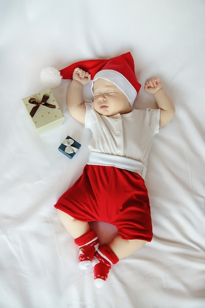 Adorável bebê recém-nascido com fantasia de Papai Noel e caixas de presente de Natal dormindo docemente em um lençol branco