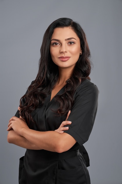 Foto adorável cosmetologista com pele clara, vestindo uniforme médico preto, posando dentro de casa. isolado sobre o fundo cinza do estúdio.