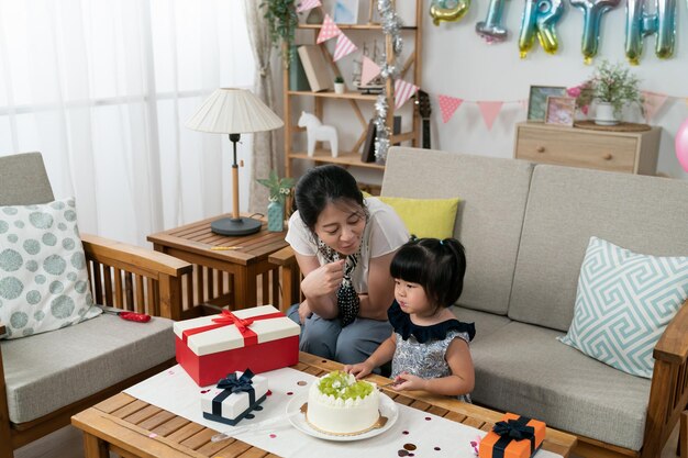 adorável criança asiática menina e mãe comendo bolo delicioso juntos em uma sala de estar brilhante com decoração de aniversário em casa