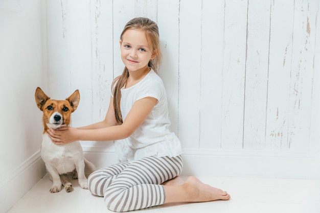 Adorável criança do sexo feminino pequena brinca com seu cachorro no quarto branco, sente-se no chão