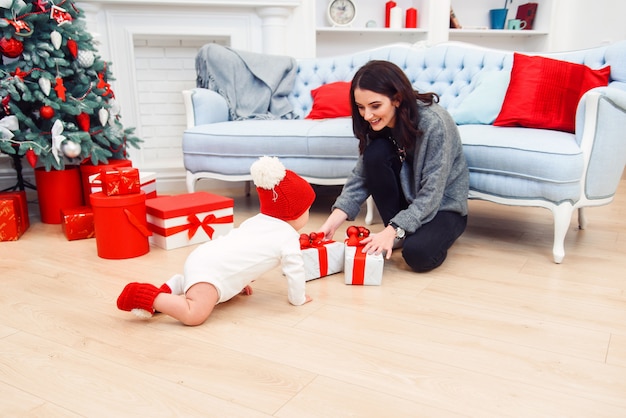 Adorável criança sorridente rastejando no parquet para os presentes de Natal perto de sua mãe, que se senta no chão.