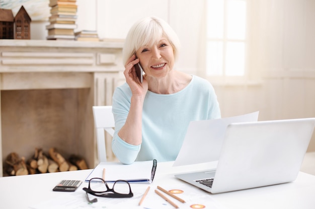 Adorável elegante mulher sênior falando com seu colega ao telefone enquanto está sentada à mesa e usando seu laptop