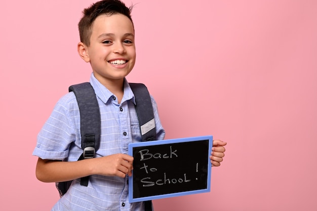 Adorável estudante com mochila segura lousa com letras de giz, volta às aulas e sorrisos fofos para a câmera, isolado sobre um fundo rosa com espaço para texto