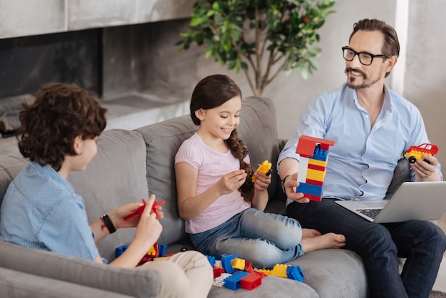 Adorável família com apenas um dos pais sentada no sofá e montando um conjunto de blocos coloridos, aparentemente se divertindo muito juntos