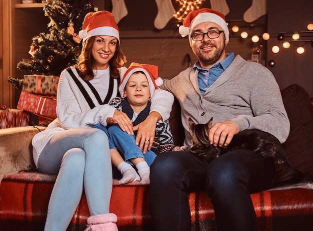 Adorável família feliz em chapéus com seu gato sentados juntos no sofá na sala decorada durante o natal.