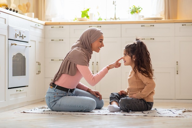 Adorável família muçulmana mãe e filha se divertindo juntos na cozinha, senhora islâmica em hijab sentada no chão e tocando o nariz de sua filha, criando laços em casa, vista lateral