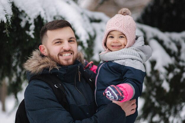 Adorável filhinha com seu adorável pai perto de uma grande árvore cheia de neve