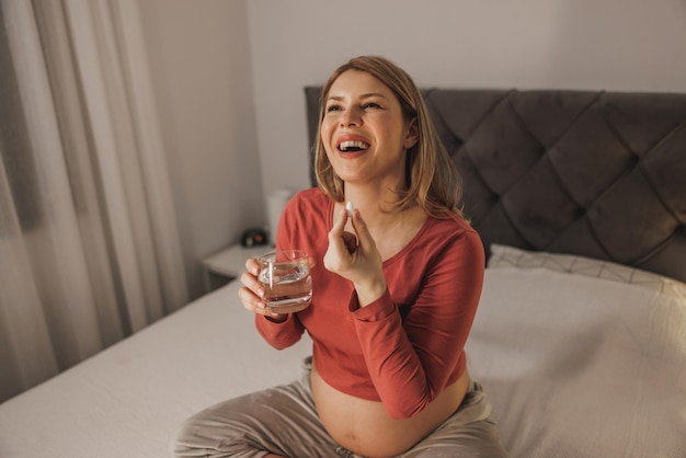 Adorável futura mãe segurando um copo de água e tomando pílulas de vitamina em uma cama no quarto.