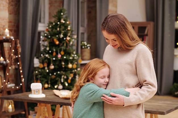 Adorável garota com cabelo ruivo comprido e ondulado dando um abraço na mãe na frente da câmera contra um pinheiro decorado e velas acesas na véspera de natal