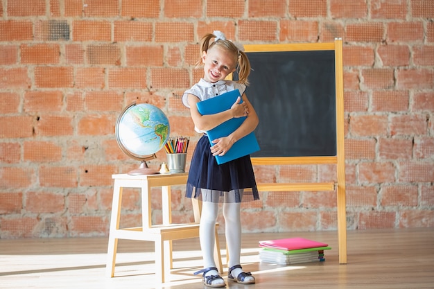 Adorável garota do jardim de infância de escola particular caucasiana sorrindo na frente do quadro-negro com um livro nas mãos, de volta ao conceito de escola