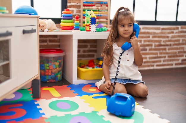 Adorável garota hispânica jogando brinquedo de telefone sentado no chão no jardim de infância