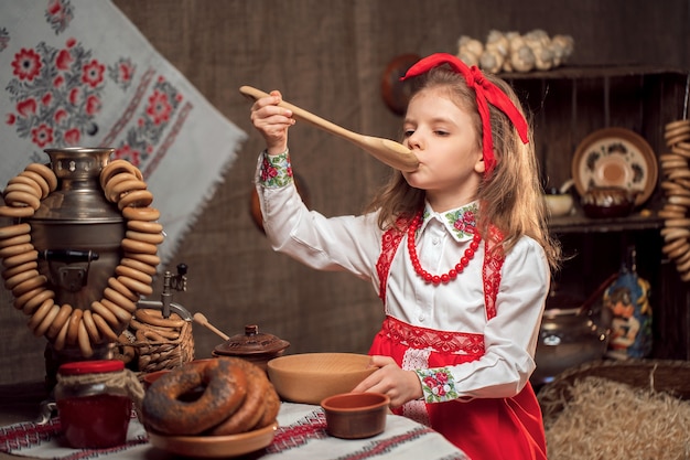 Adorável garota sentada à mesa cheia de comida e grande samovar. Maslenitsa tradicional de comemoração