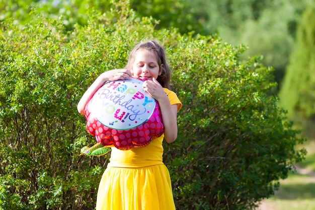 Adorável garota vestida de amarelo com balão colorido de feliz aniversário festivo em forma de flor. Criança feliz ao ar livre