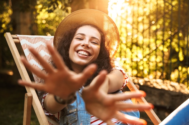 Adorável jovem com cabelo castanho encaracolado usando chapéu de palha, sorrindo e descansando no parque enquanto está sentada na espreguiçadeira durante o fim de semana ensolarado