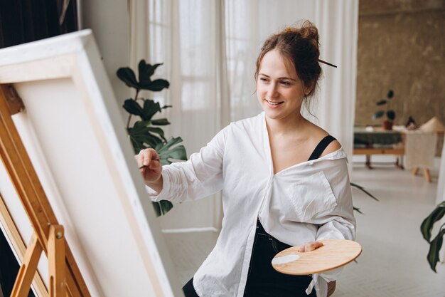 Adorável jovem com roupa casual criando desenhos no cavalete no estúdio brilhante