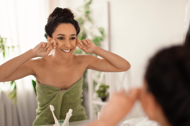 Adorável jovem fazendo auto-massagem facial de drenagem linfática perto do espelho em casa espaço livre